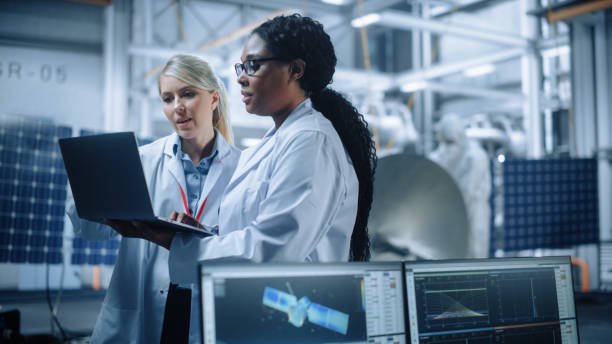 Two Female Engineers Talk, Use Computer working at Aerospace Satellite Manufacturing Facility. Diverse Team of Multi-Ethnic Scientists Doing Science Technology Research for Space Exploration Program
