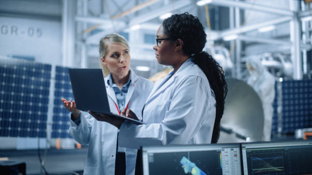 istock Two Female Engineers Talk, Use Computer working at Aerospace Satellite Manufacturing Facility. Diverse Team of Multi-Ethnic Scientists Doing Science Technology Research for Space Exploration Program 1323369485