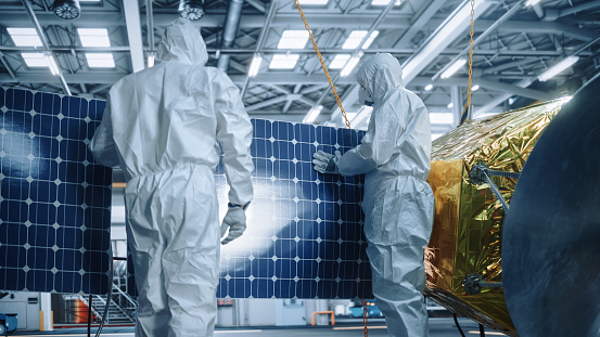 Engineer and Technician in Protective Suits Working on Satellite Construction. Aerospace Agency: Team of Scientists Fixing Solar Panel Wings to Spacecraft. International Space Exploration Mission