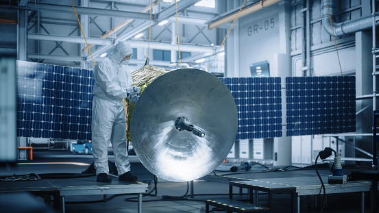 Engineer in Protective Suit Working on Satellite Construction. Aerospace Agency Manufacturing Facility: Scientists Developing Spacecraft for Space Exploration, Communications, Cosmos Observation