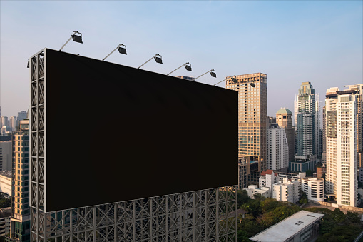 Blank black road billboard with Bangkok cityscape background at sunset. Street advertising poster, mock up. Side view. The concept of marketing communication to promote or sell idea.