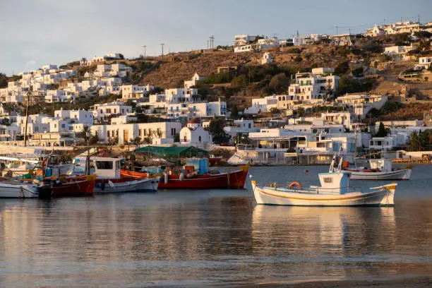 Greek fishing boats in Mykonos island port, whitewashed buildings, cityscape background. Cyclades, Greece. Colorful traditional boats anchored on calm sea water at sunset, reflections