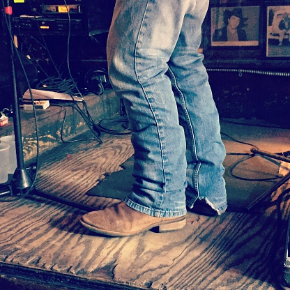 Close up of cowboy boots and blue jeans legs on well worn stage with lower part of microphone stand and wires.