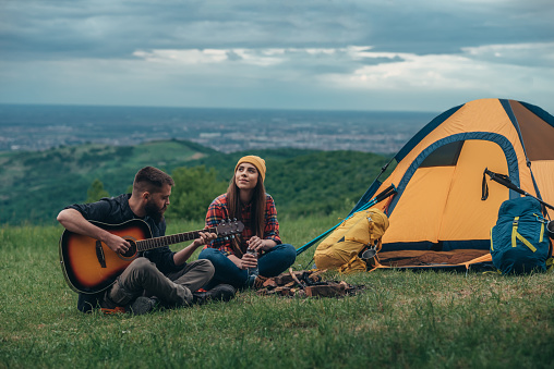 Young couple of campers sitting next to the tent and playing guitar while having fun in the nature