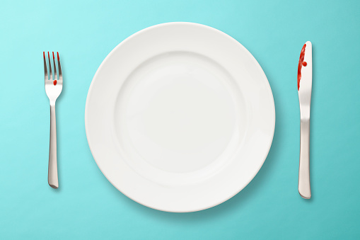 Overhead shot of empty plate and knife and fork with ketchup on them, on light blue background.
