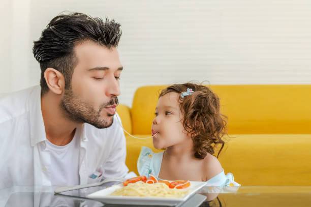 An absolutely adorable and attractive daddy and daughter are having some delicious with spagetti together at home stock photo