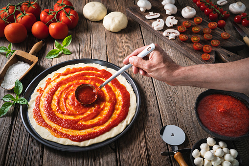 Italian pizza male hand spreading tomato sauce with ladle on the pizza raw dough with ingredients as mozzarella, mushrooms and tomato on rustic wooden table