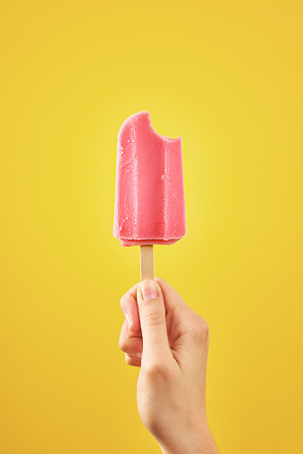 Woman holds bitten red ice cream on yellow background. Color frozen fruit popsicle.