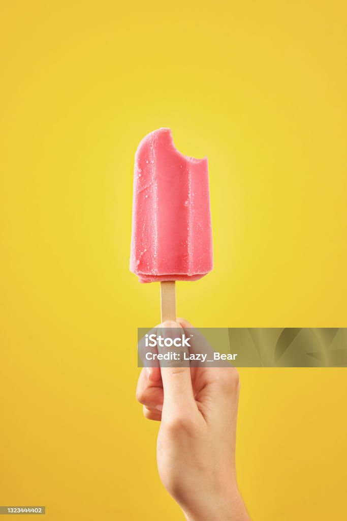 Bitten red frozen fruit ice cream popsicle on yellow background Woman holds bitten red ice cream on yellow background. Color frozen fruit popsicle. Flavored Ice Stock Photo