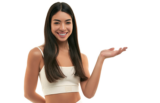 Young Asian woman with clean radiant healthy skin holds on the palm, shows on the product isolated on white background. Facial skin care concept, cosmetology, spa.