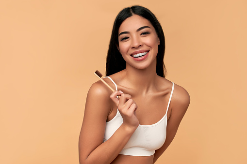 Young Asian woman in white lingerie with a toothbrush and white teeth on a beige background. Dental hygiene concept. Dentistry.