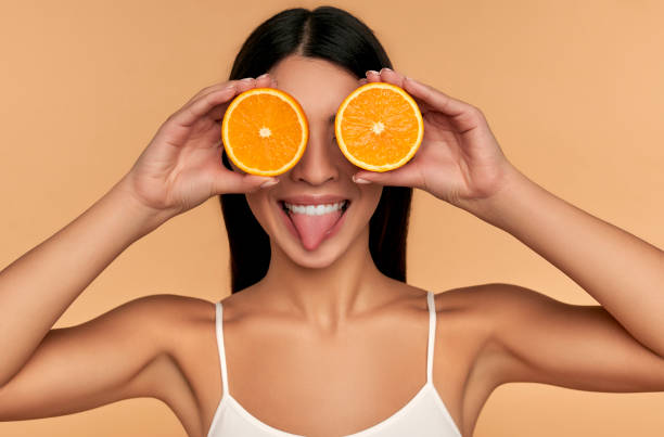 Beauty woman on background Portrait of Asian girl with shining clean skin of face holding orange halves in white underwear isolated on beige background. Vitamin C cosmetics concept oranges with faces stock pictures, royalty-free photos & images