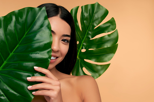 Young Asian woman with clean radiant face skin with green leaf on beige background. Spa care, facial skin care, beauty cosmetology. Natural cosmetics concept.