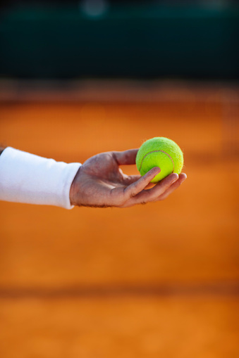 An unrecognisable persons hand holding a tennis ball on a clay court