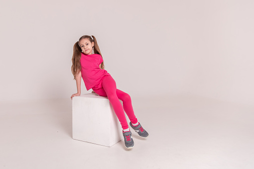 beautiful sports girl in a pink T-shirt and pants on a white isolated background. child sitting on a white cube. girl with tails. Copy space
