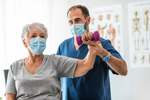 Senior emale patient in a medical mask exercising with dumbbell in for rehabilitation exercises, while is being assisted for a physiotherapist or doctor, in a hospital or clinic. Covid19 outbreak.