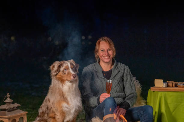 istock Young woman drinks red wine by the campfire in the forest. Her Australian Shepherd sits next to her on a rug in the snowy grass. Blue smoke curls up in the dark 1323459206