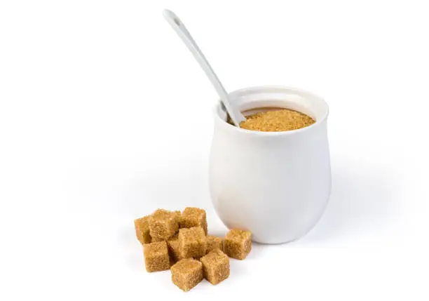 Natural unrefined brown granulated sugar in white sugar-bowl with spoon and cubes of brown sugar scattered beside on a white background