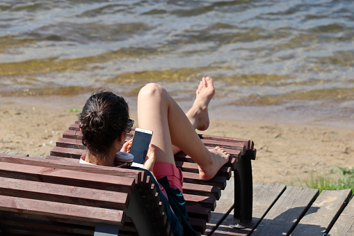 Moscow, Russia - June 2021: Girl sitting on deck chair with smartphone on a beach of Moscow river in Serebryany Bor