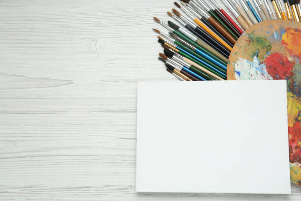 istock Blank canvas, brushes and palette on white wooden table, flat lay. Space for text 1323473351