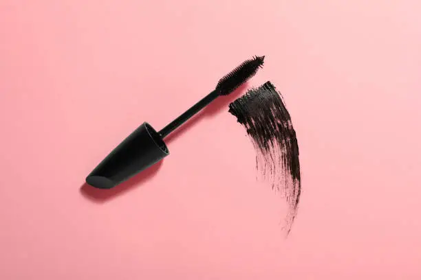 Mascara wand and smear on pink background, top view. Makeup product