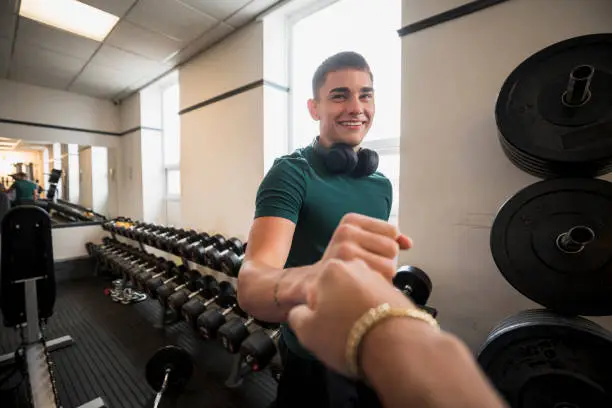 A point of view shot of an unrecognisable mixed race man fist bumping his friend in the gym. His friend is wearing sports clothing and has wireless headphones around his neck.