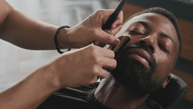 4k video footage of a handsome young man sitting in a barbershop and getting his beard shaved by a barber