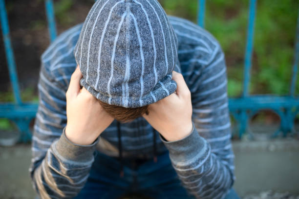 A lonely and sad teenage student in a hood covered his face with his hands, hiding from problems on the street in an urban environment. stock photo