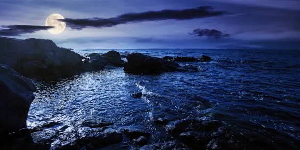 sea coast scenery at night. boulders in the calm water. few clouds on the sky in full moon light. lonely place for summer vacations. sunny weather