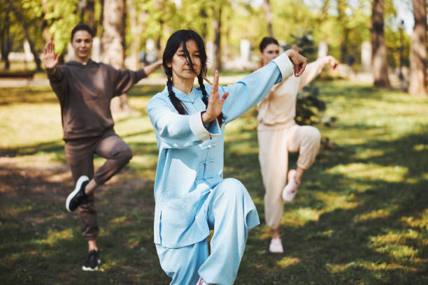 Tai chi trainer standing in crane pose before two students Woman martial arts master staying on right leg outdoors while showing her palm to camera with pair of students woman legs open stock pictures, royalty-free photos & images