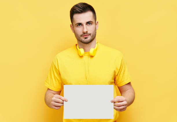 Young caucasian man holding white blank card with copy space for text on yellow background