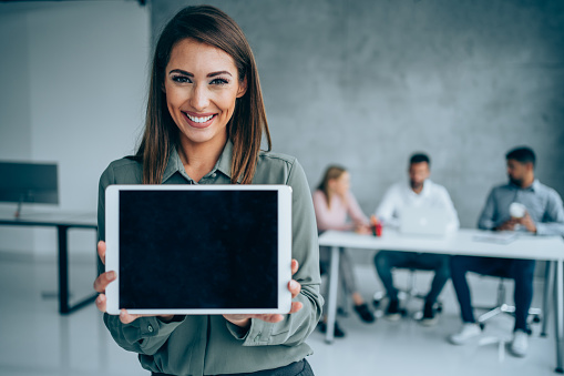 Beautiful young businesswoman standing in the office with her team in background and showing black empty screen with copy space on digital tablet.