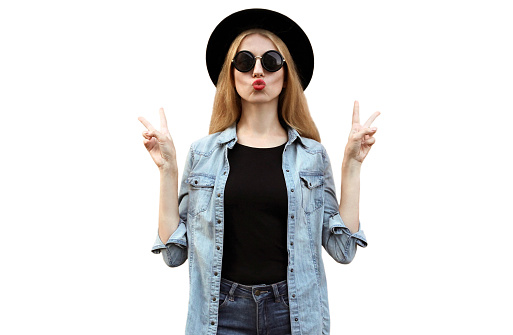 Portrait of stylish young woman blowing her lips with red lipstick wearing a black round hat, jeans jacket, female model posing isolated on a white background