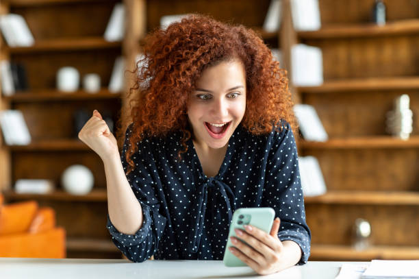 istock Surprised curly red-haired young woman using smartphone indoors 1323520608