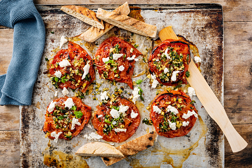 Overhead view of thick slices of beefsteak or heirloom tomatoes cooked with; garlic, ginger, coriander and served with fresh arugula, mozzarella cheese and slices of pitta bread. Horizontal format, with some copy space photographed against a rustic wooden background.