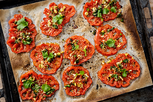 Overhead view of thick slices of beefsteak or heirloom tomatoes cooked with; garlic, ginger, coriander and sesame seeds. Horizontal format, with some copy space photographed on a baking tray just out of the oven.