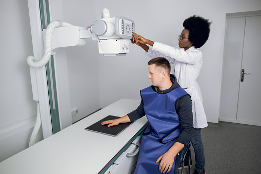 Classic ceiling-mounted x-ray system. Young African woman doctor radiologist and male Caucasian patient in x-ray room, during X-ray of human hand. Xray diagnostics of bone fracture