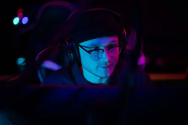 Photo of A young esports player sits at a computer during an online game tournament. Neon lighting of the esports arena. A hat and headphones with a headset