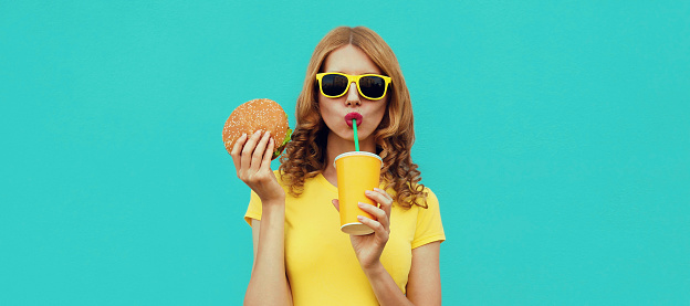 Portrait close up of young woman with fast food, burger and cup of juice wearing a yellow t-shirt, sunglasses on a blue background