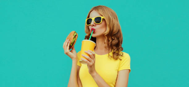 porträt nahaufnahme der jungen frau mit fast food, burger und tasse saft mit einem gelben t-shirt, sonnenbrille auf blauem hintergrund - freedom sandwich bread food stock-fotos und bilder