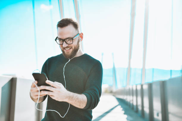 istock Smiling Bearded Male Setting Up Playlist On Smartphone While Walking With Headphones On Bridge 1323549095