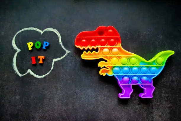 Photo of Pop it dinosaur toy rainbow colors on a black background with multicolored letters and the inscription - Pop it in a speech bubble