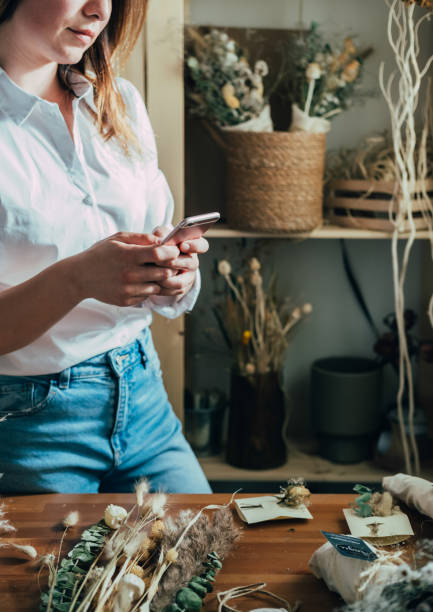 istock Unrecognizable Smiling Woman Entrepreneur Using Mobile Phone in her Workshop Full With Different Dry Flowers and Herbs 1323555654