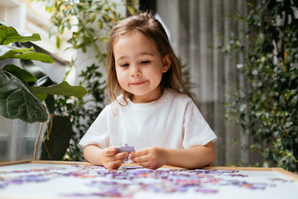 Little girl is trying to connect couple jigsaw puzzle piece. Side view of concentrated little girl playing with puzzles at home