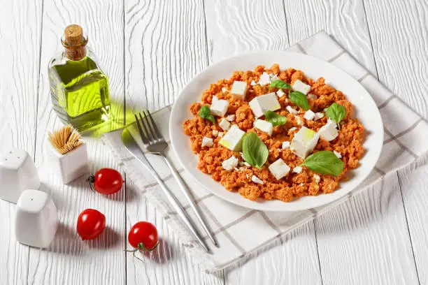 Strapatsada, eggs scrambled with tomatoes and feta cheese on a white plate with fresh basil leaves, horizontal view from above, greek recipe, close-up