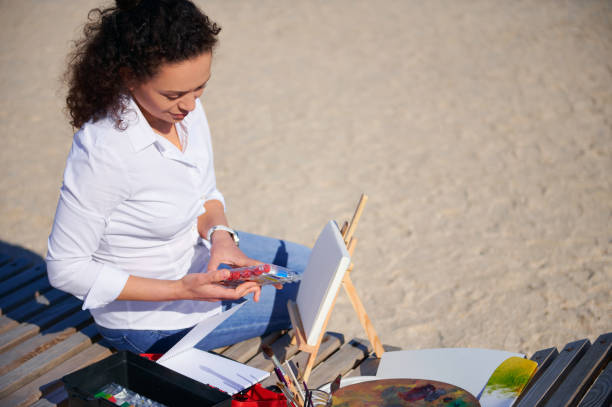 istock Young painter opening a box with paints ready to paint on canvas in open air sitting on a wooden chaise lounge on the beach 1323559210