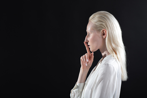 Closeup of beautiful young white caucasian albino blond woman with finger on her lips showing shhh silence gesture on black background.