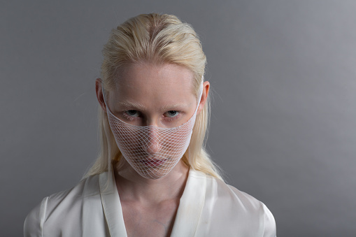 Closeup portrait of white caucasian albino blond woman fashion model wearing quarantine medical face mask coarse mesh net on grey background. Covid-19 coronavirus protection concept.