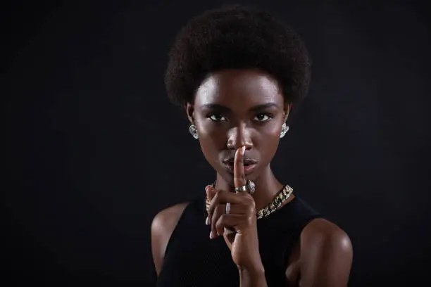 Photo of Closeup of beautiful young dark-skinned woman with finger on her lips showing shhh silence gesture on black background.