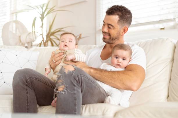 Handsome new father enjoys time with his twin sons A handsome young father of hispanic descent is sitting on a white leather couch. He is holding both his newborn fraternal twin baby sons, one in each arm. He is looking down at one of them and smiling. He is a proud new dad. twin photos stock pictures, royalty-free photos & images